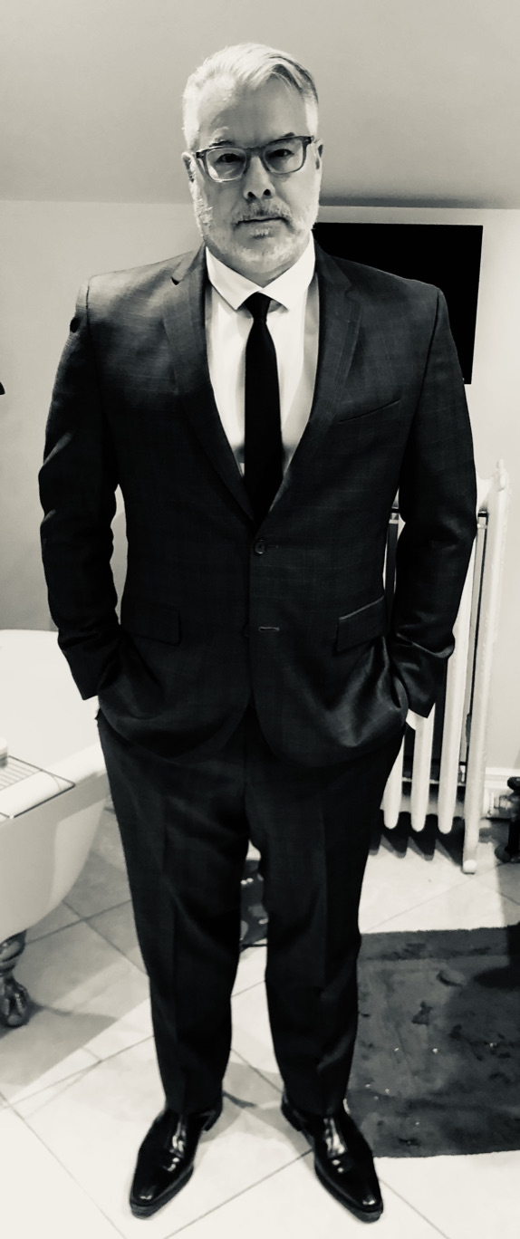 A black and white picture of Mark in a suit, hands in pocket, getting ready to go somehwere